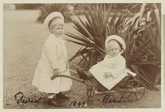 Photograph of Prince Edward of York (1894-1972), later the Duke of Windsor, standing in three-quarters right whilst holding the arms of a wheelbarrow in which Prince Albert of York (1895-1952), later King George VI, is sitting. Both children wear sailors hats with a band around the rim that bears the name 'Britannia', the Royal Cutter built in ... Royal Collection Trust/© Her Majesty Queen Elizabeth II 2014